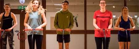 Five people hold red-handled scooters, standing side by side and facing forward, indoors in a brightly lit room with glass-panel doors and a green exit sign in the background.