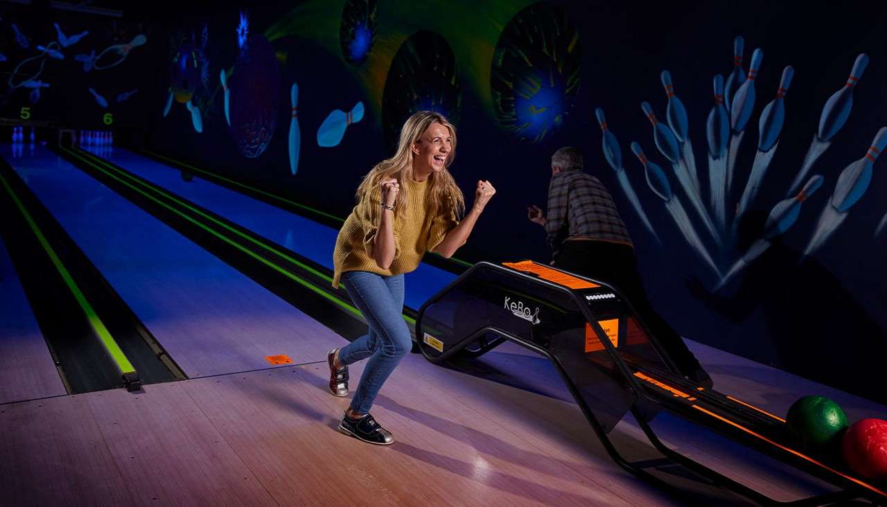 Woman celebrates, fists raised, after bowling; behind her, another person uses a ramp. Cosmic-themed glow-in-the-dark lanes and pin murals surround; lane numbers 5 and 6; ramp labeled KeBoL.
