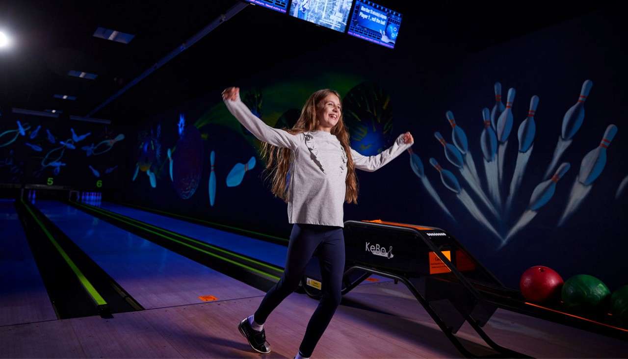 Child raises arms celebrating after bowling, near ball return beside neon-lit lanes. Murals of pins decorate walls; colored balls sit nearby. Visible text: “KeBoL” on ball return, “5” at lane end.