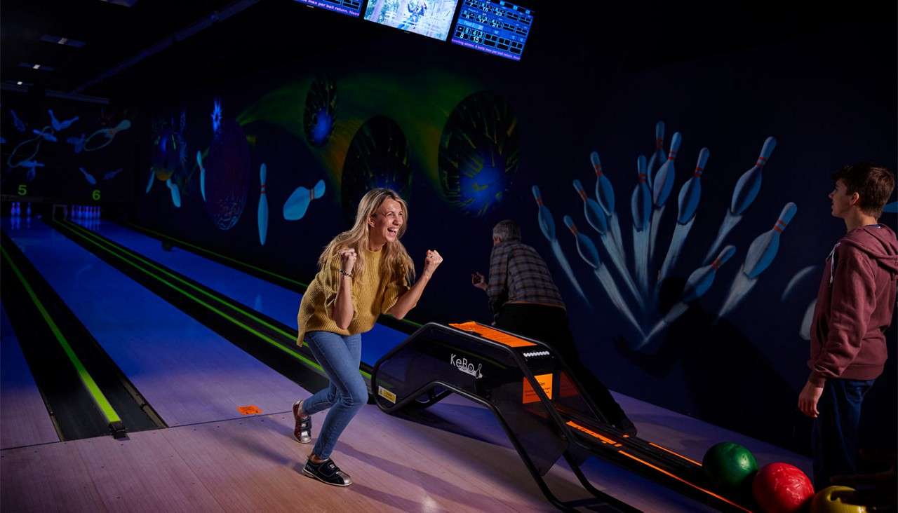 Woman pumps her fists after bowling beside a ball ramp labeled “KeBoL”; companions watch. Neon-lit bowling alley with glowing lanes, pin murals, and lane numbers “5” and “6.”