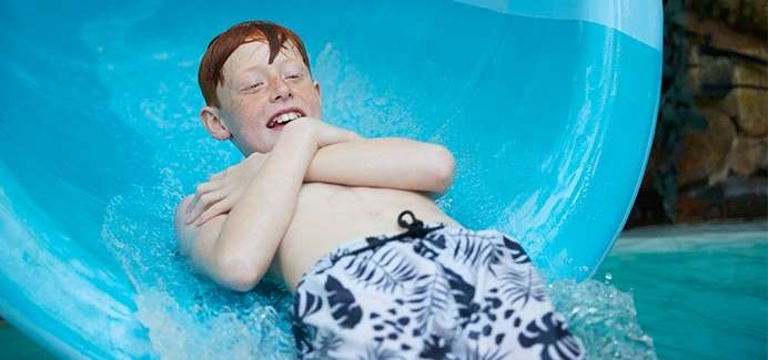 Boy slides down blue tube waterslide, arms crossed and smiling, water splashing around him, wearing black-and-white leaf-print swim trunks; indoor waterpark setting with rock wall in background.