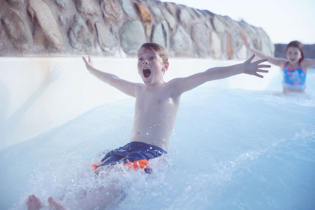 Child with arms outstretched slides through shallow water, splashing and shouting with excitement; another child follows behind. Context: outdoor water park channel bordered by a stone wall under bright light.