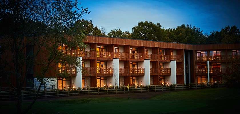 Multistory lodge glows with warm balcony lights, columns punctuating its facade. At dusk, it overlooks a manicured lawn with a golf flag, bordered by wooden fence and surrounding trees.