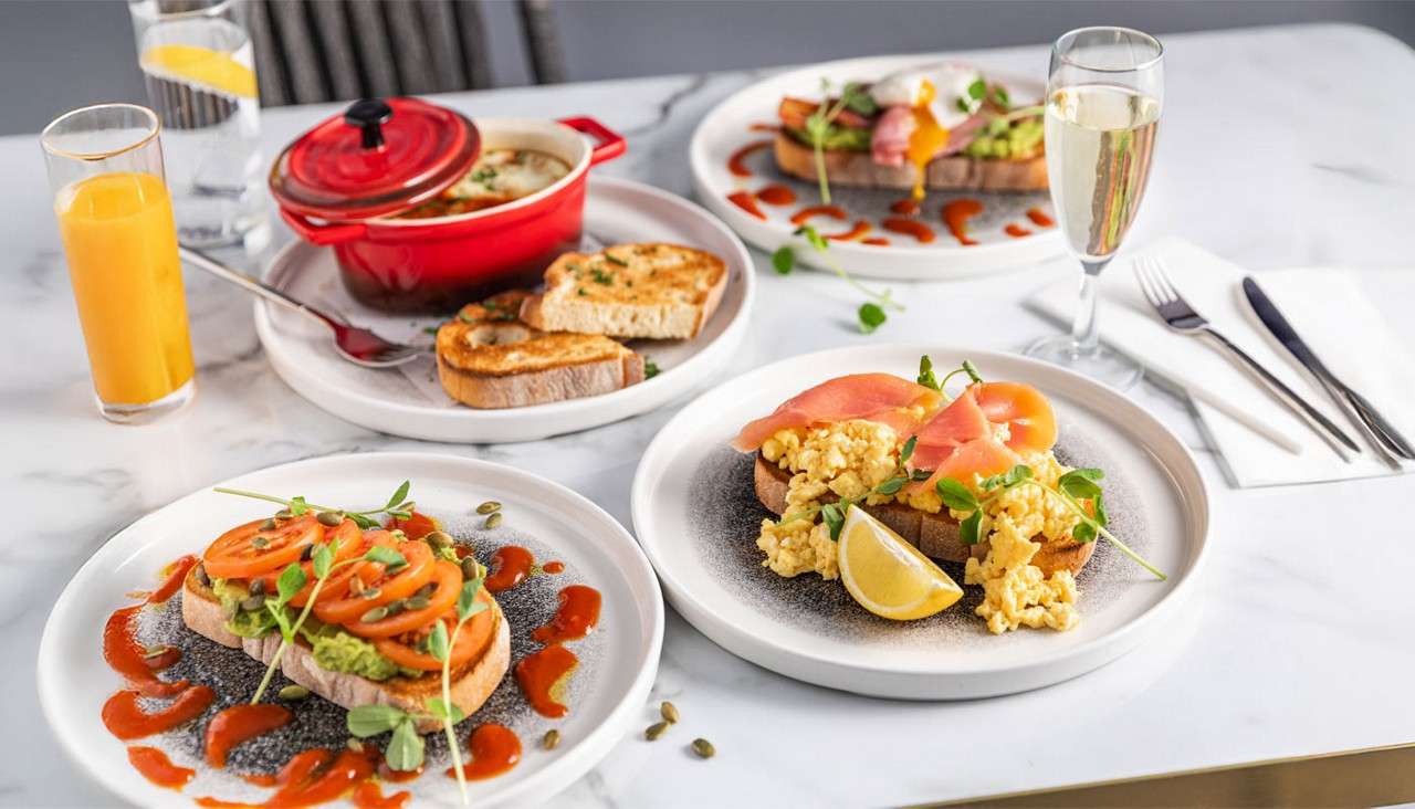 Brunch plates arranged: avocado-tomato toast with sauce; scrambled eggs and smoked salmon with lemon; a red pot of soup with toast; plus orange juice and wine on a marble table.