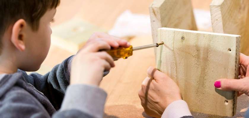 Child with a screwdriver tightens a screw while an adult holds two wooden boards steady on a workbench, suggesting a collaborative woodworking project in a bright indoor setting.
