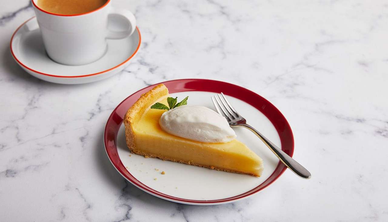 Slice of lemon tart topped with whipped cream and mint sits with a fork on a red-rimmed plate; a cup of coffee on a saucer rests on a marble tabletop.