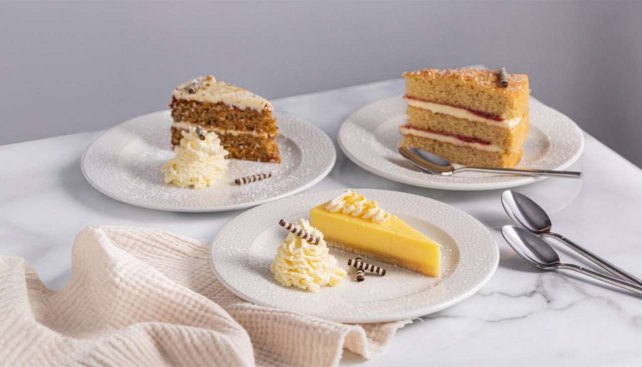 Three plated desserts sit on a marble table: carrot cake, layered sponge with cream and jam, and lemon tart; each dusted with sugar beside whipped cream, spoons, and beige cloth.