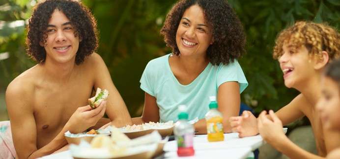 Group of people smile and eat together, chatting. They sit at a picnic table with bowls, bread, and small juice bottles. Sunlit greenery surrounds them, suggesting a relaxed outdoor gathering.