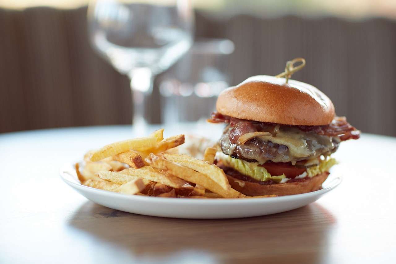 Cheeseburger with bacon and melted cheese rests on a white plate beside golden fries, placed on a wooden table in a softly lit indoor setting.