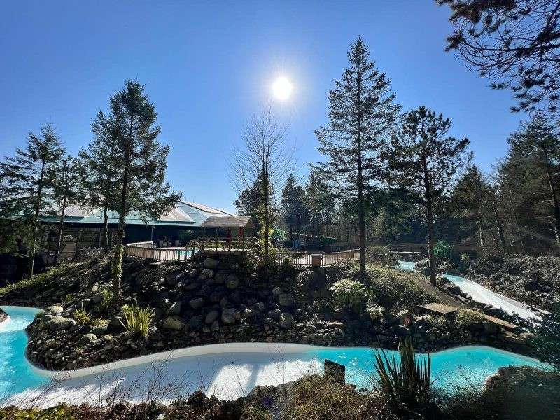 Lazy river winds through a rocky landscaped area, sunlit turquoise water flowing beneath tall evergreens. A low bridge and building sit nearby under a clear blue sky.