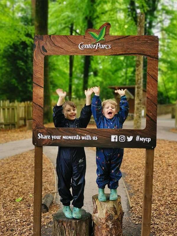 Two children raise their arms and smile, standing on tree stumps within a wooden photo frame in a lush forest path. Text: CenterParcs; Share your moments with us; Facebook Instagram Twitter icons; #mycp.