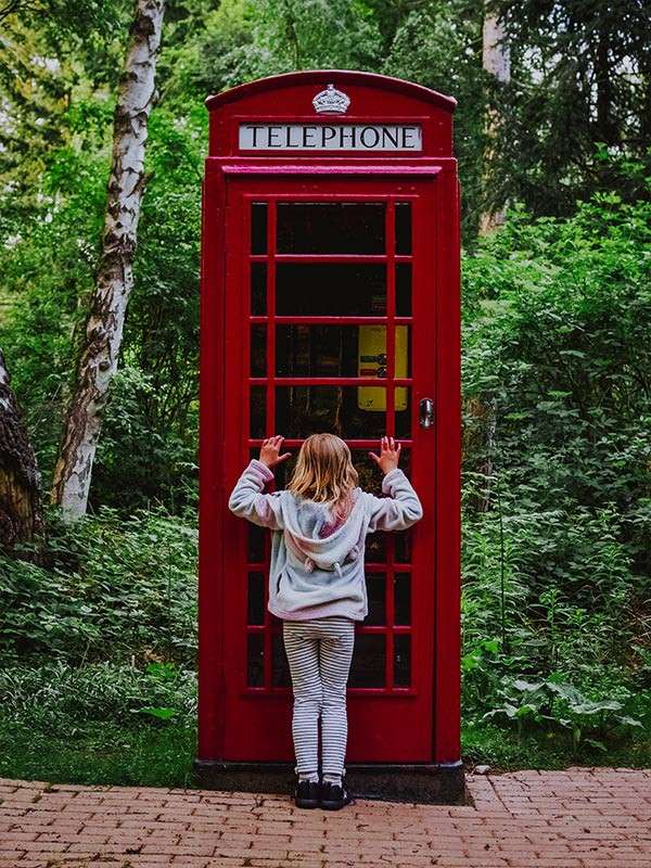 Child presses hands to a red phone booth door, peering inside; the sign reads TELEPHONE, and the booth stands on a brick path amid dense green forest.