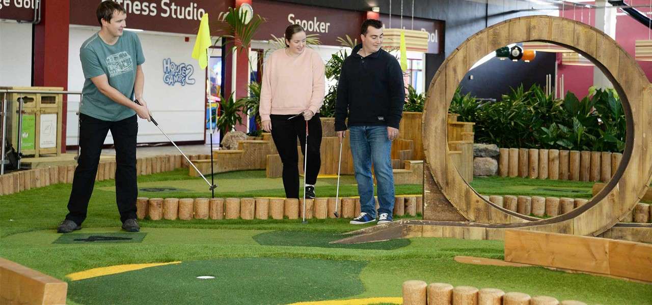 Three people putt at an indoor mini-golf course, one mid-swing, beside a large wooden loop obstacle amid plants and turf. Text: Fitness Studio; Snooker; Pool; House of Games.