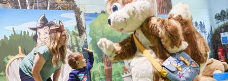 Large squirrel mascot in a sling bag high-fives a small child while a smiling adult watches nearby, in an indoor event space decorated with forest-themed murals and props.