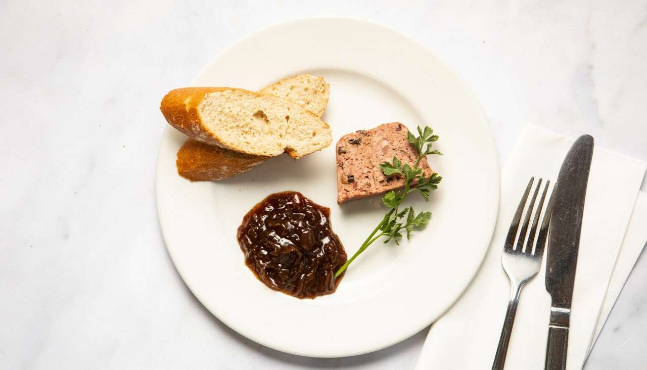 A small appetizer sits arranged: baguette slices and a pâté square with parsley accompany glossy onion chutney; beside the plate, a knife and fork rest on a napkin on marble.