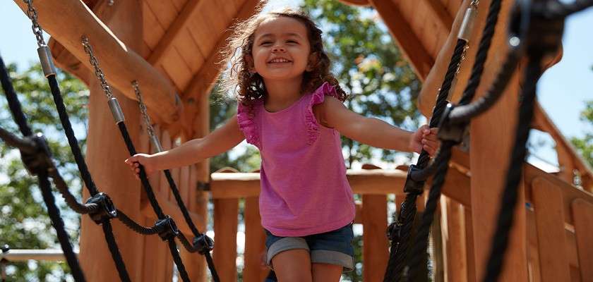 Child in a pink shirt walks across a rope bridge, gripping black ropes, smiling; context: wooden playground structure under trees on a sunny day.