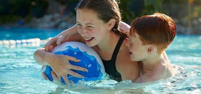 Two children embrace a blue-and-white beach ball, laughing as they play in waist-deep pool water; sunlight glints on ripples, lane rope and greenery blur in the background.
