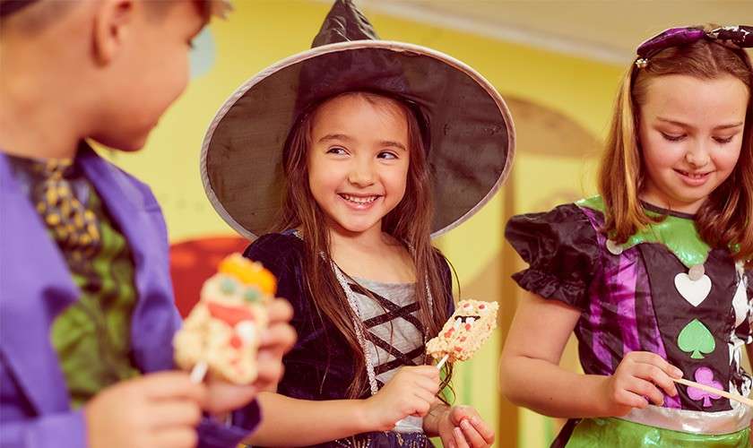 Three children in Halloween costumes share decorated treats; the center girl, dressed as a witch, smiles while holding a rice-krispie pop; classmates beside her examine theirs in a brightly lit classroom party.