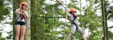 Two harnessed children traverse suspended ropes; one balances on a vertical rope line, the other grips safety lines, in a forest treetop adventure course among tall trees.