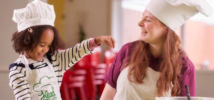 Child sprinkles ingredients while a smiling chef supervises, both wearing white hats and aprons, in a bright kitchen classroom with blurred red chairs and utensils in the background.