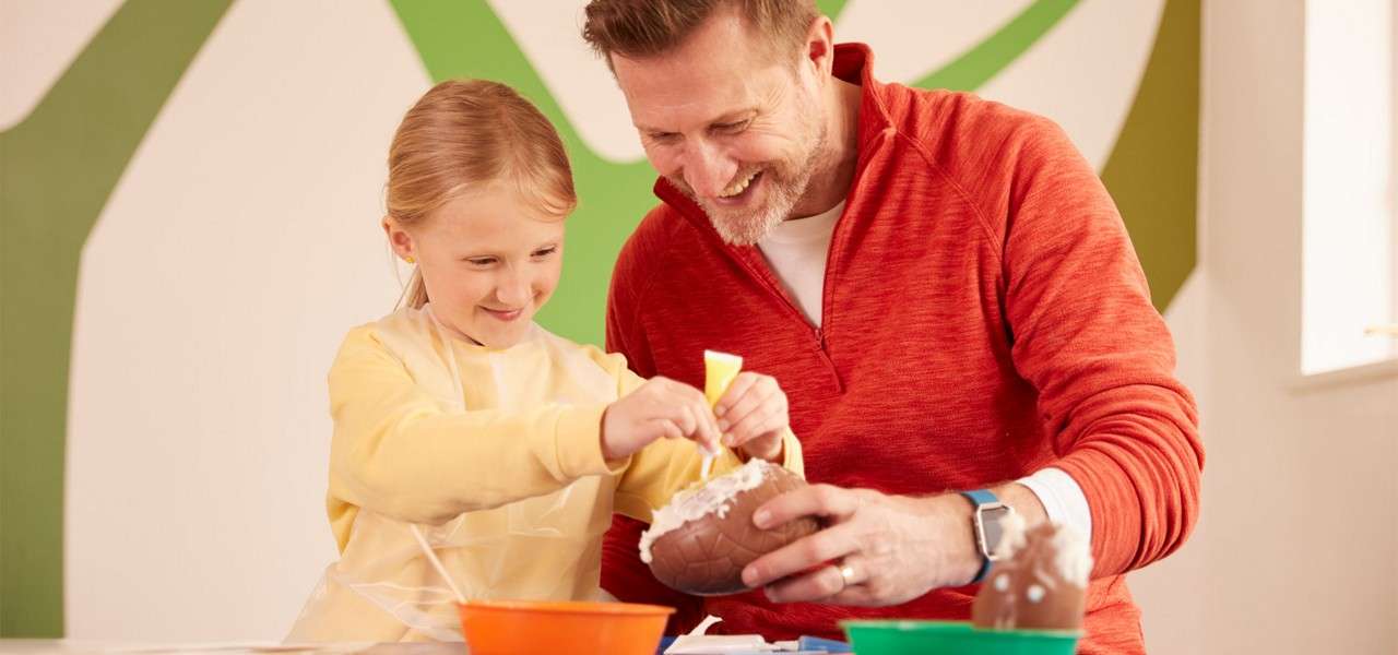Chocolate egg receives white icing as an adult and child squeeze a piping tube, working at a table with colorful bowls in a bright room with abstract green wall shapes.