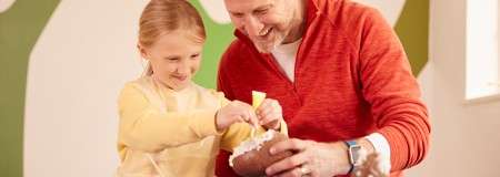 Child and adult spread icing onto a large chocolate egg, smiling together at a table in a bright indoor craft space with soft green wall shapes.