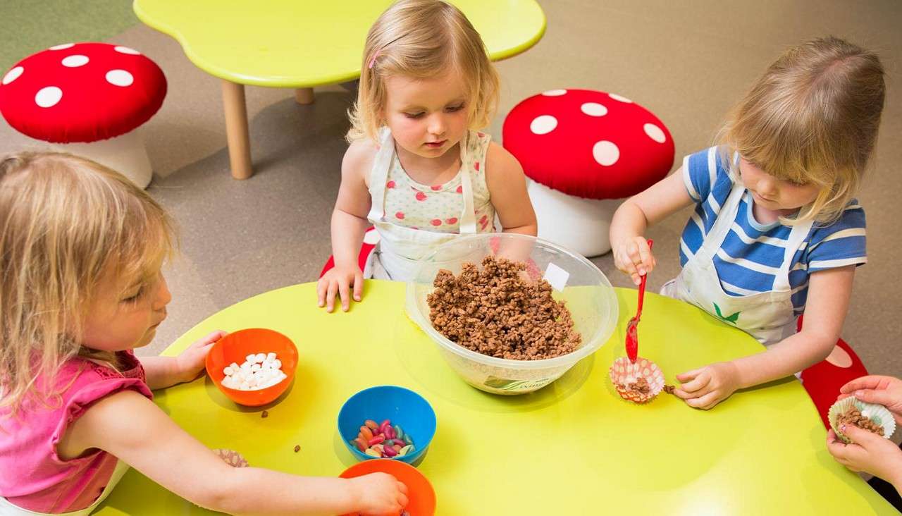 Children spoon chocolate cereal mixture into paper cupcake cases, seated around a bright yellow round table. Bowls of marshmallows and jelly beans surround them, with mushroom-pattern stools in a playroom.