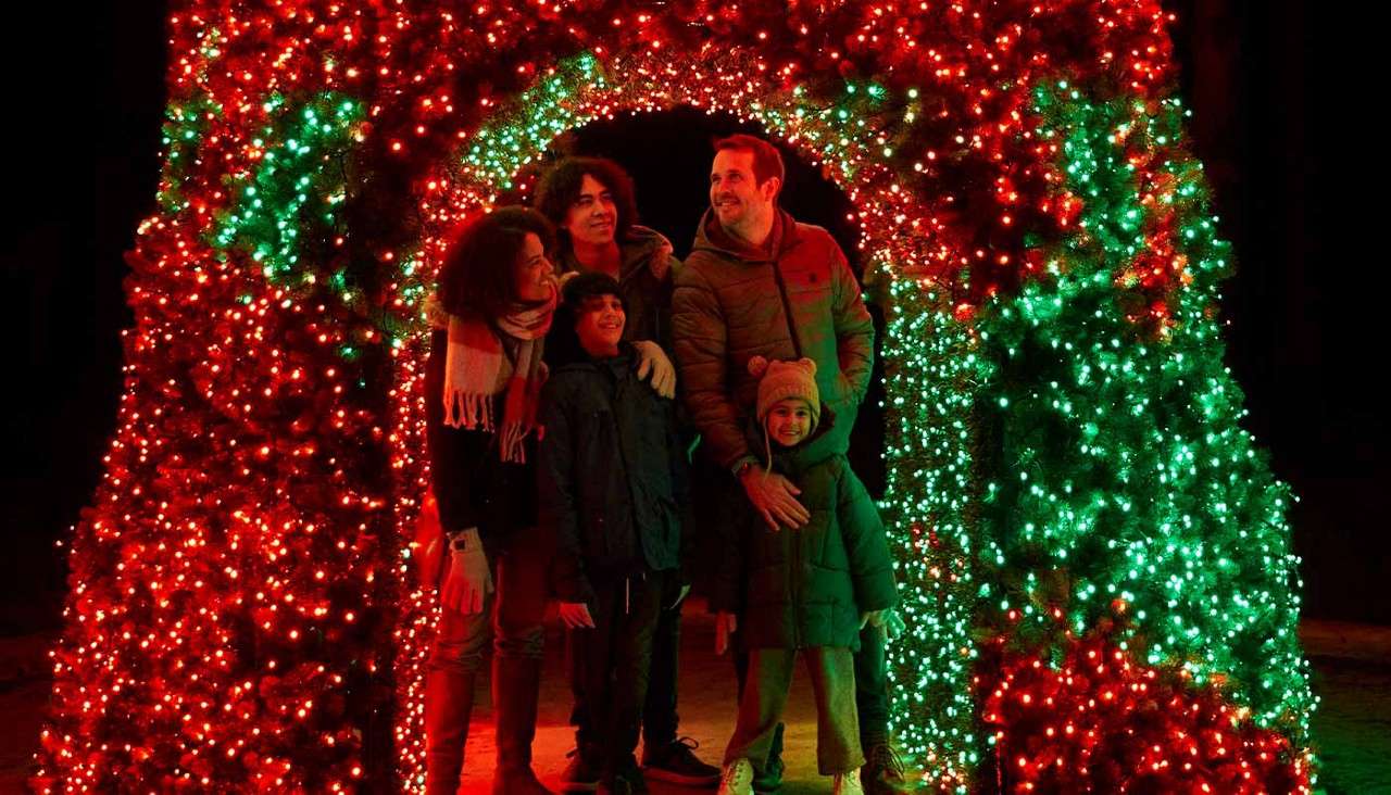 Group of five people stand smiling, bundled in winter coats, posing under a tunnel of red and green holiday lights, at night in a festive outdoor display.