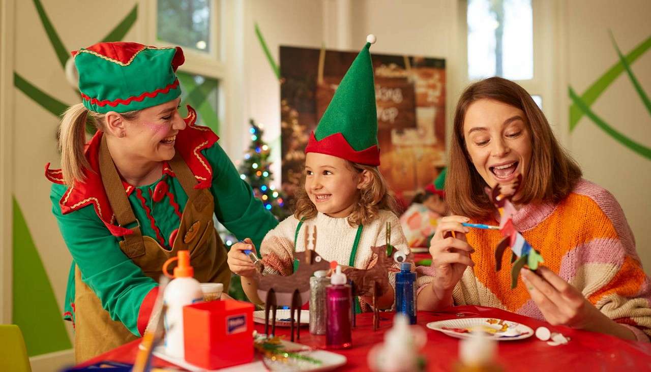 Child paints cardboard reindeer while smiling between two adults in elf outfits; they craft with glue, glitter, and brushes at a red table in a festive room with Christmas tree.