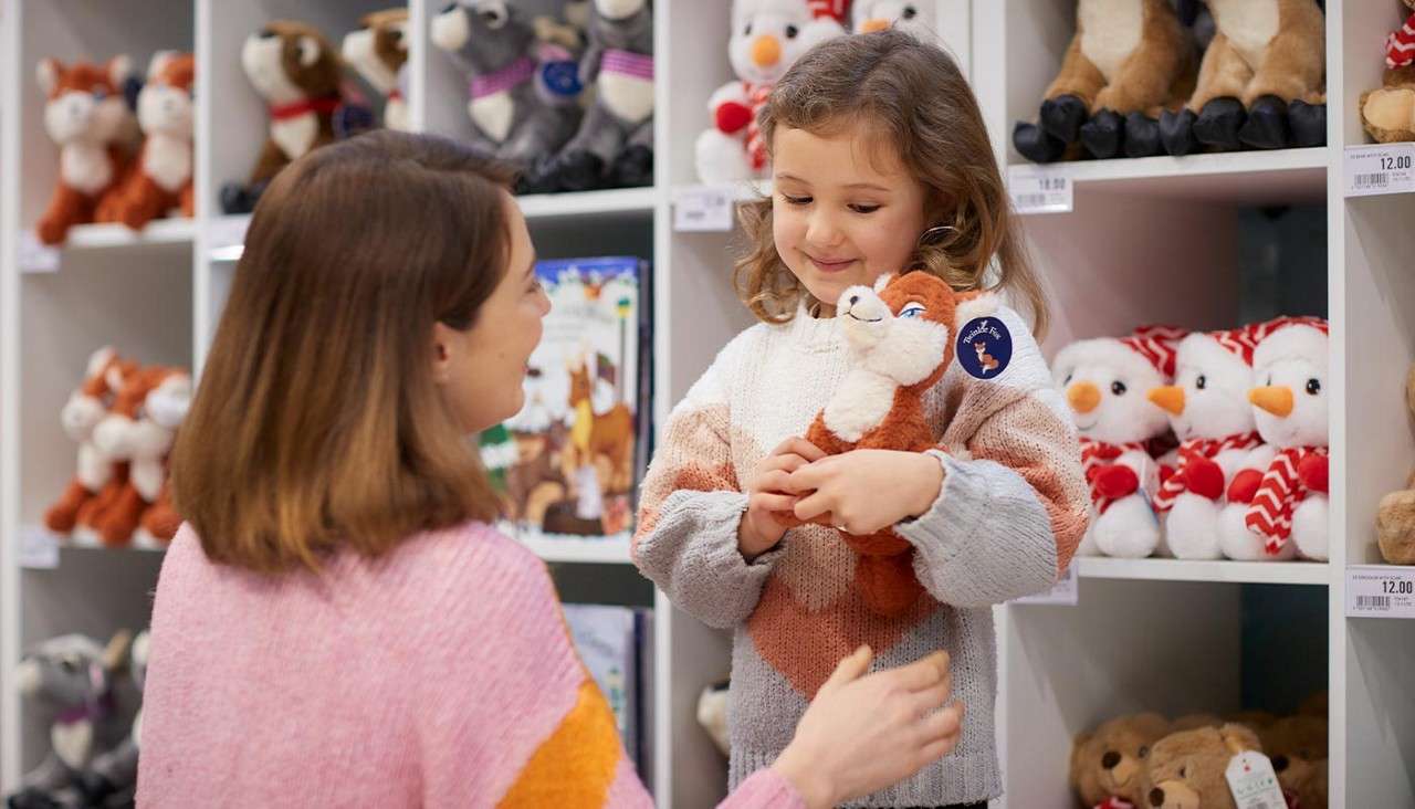 Child hugs a plush fox while smiling at an adult, in a toy store with shelves of stuffed animals; visible shelf labels say: 12.00.