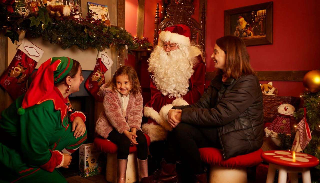 Child smiles and chats with Santa while an elf and a woman look on, seated in a cozy Christmas room with stockings, garlands, and a tree near a fireplace.