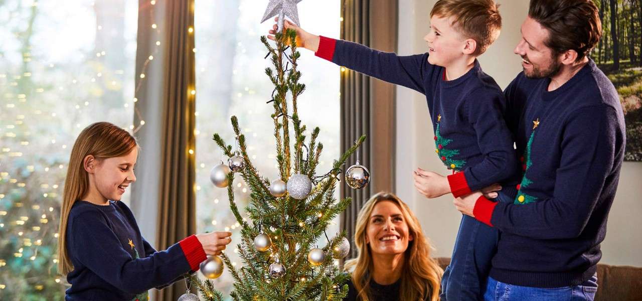Christmas tree being decorated with silver baubles; an adult lifts a child to place the star, while another child and adult assist, inside a lit living room with fairy lights.