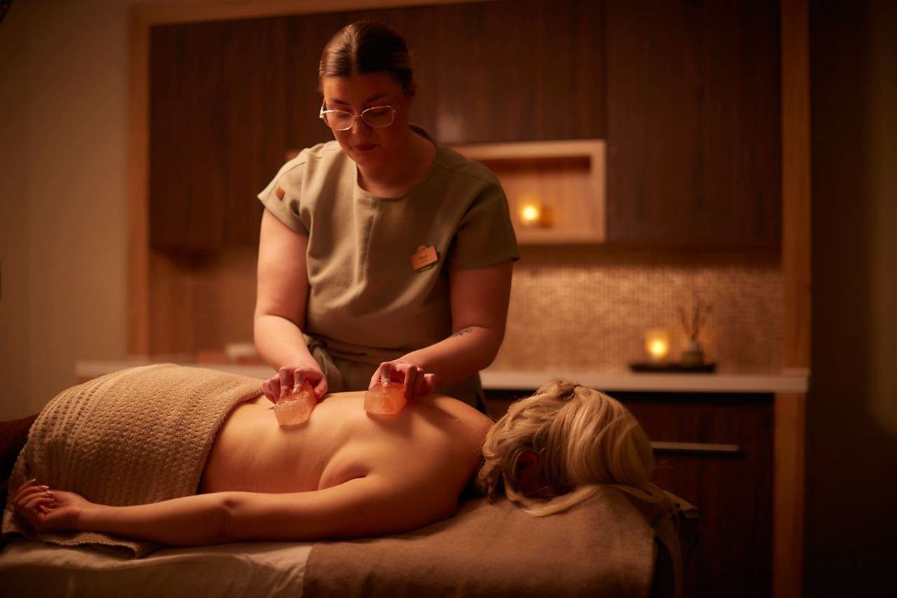 Massage therapist applies two pink salt stones to a client’s back in a dimly lit spa room with candles, wooden cabinets, and a towel-covered massage table.