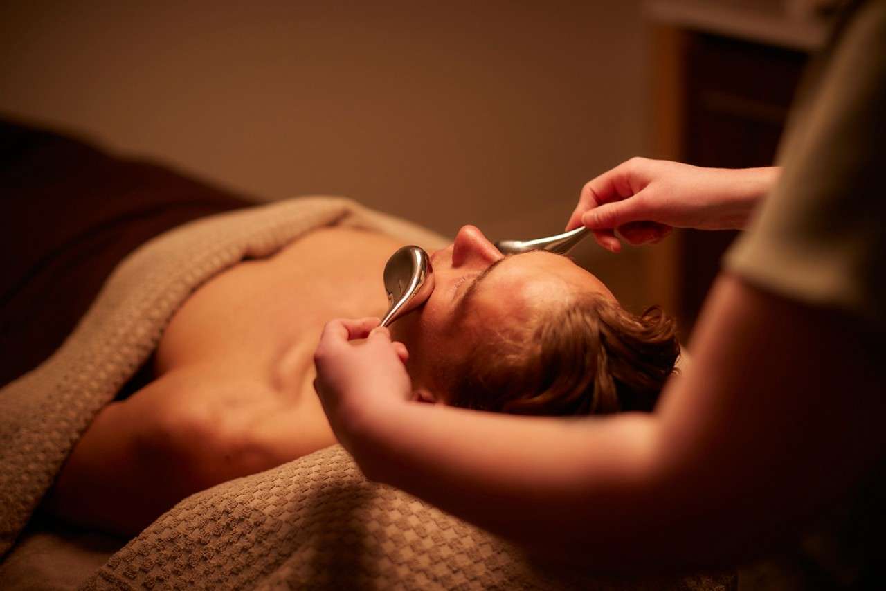 Client receiving treatment as therapist glides two metal spoons over closed eyes and forehead, on a massage table under a textured beige blanket in warm, dim spa lighting.