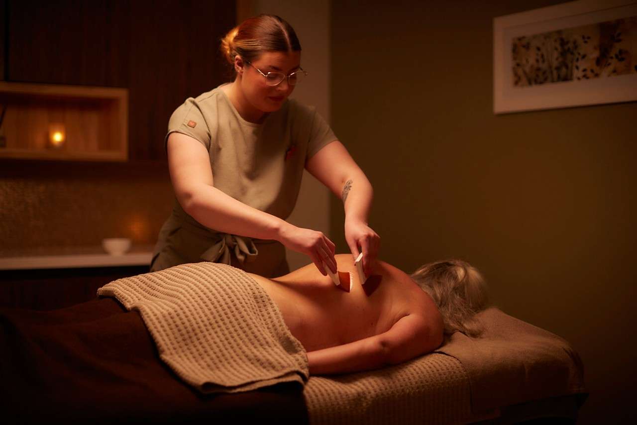 Massage therapist uses two flat tools to scrape a client’s bare back in a dimly lit spa room with warm lighting, a candle, textured blanket, and wall art.