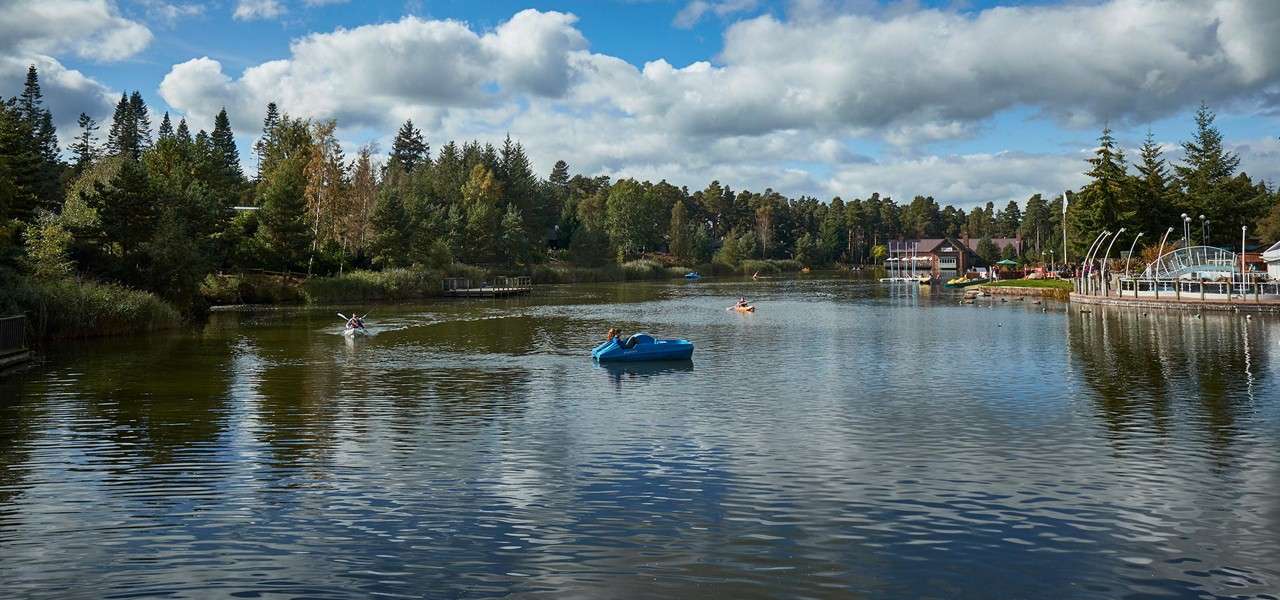 Paddleboats and kayaks glide across a calm lake, creating ripples, surrounded by trees and lakeside buildings, with a dock and curved structures on the right under a cloudy sky.