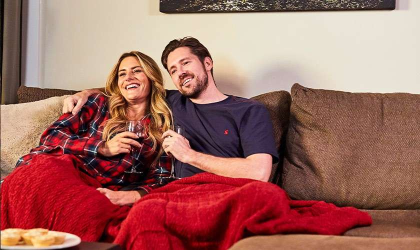 Two adults sit close together, smiling and holding wine glasses, lounging under red blankets. They relax on a brown sectional sofa in a cozy living room with snacks nearby.