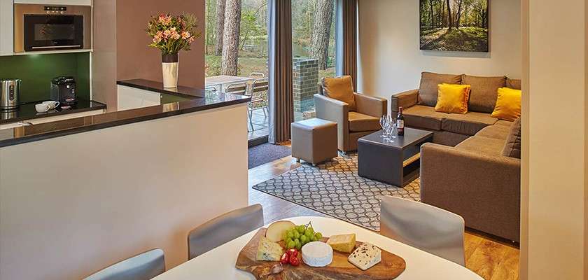 An L-shaped sofa anchors the space, facing a coffee table with wine; nearby, a dining table displays a cheese board; beyond, sliding doors open to a patio and trees.