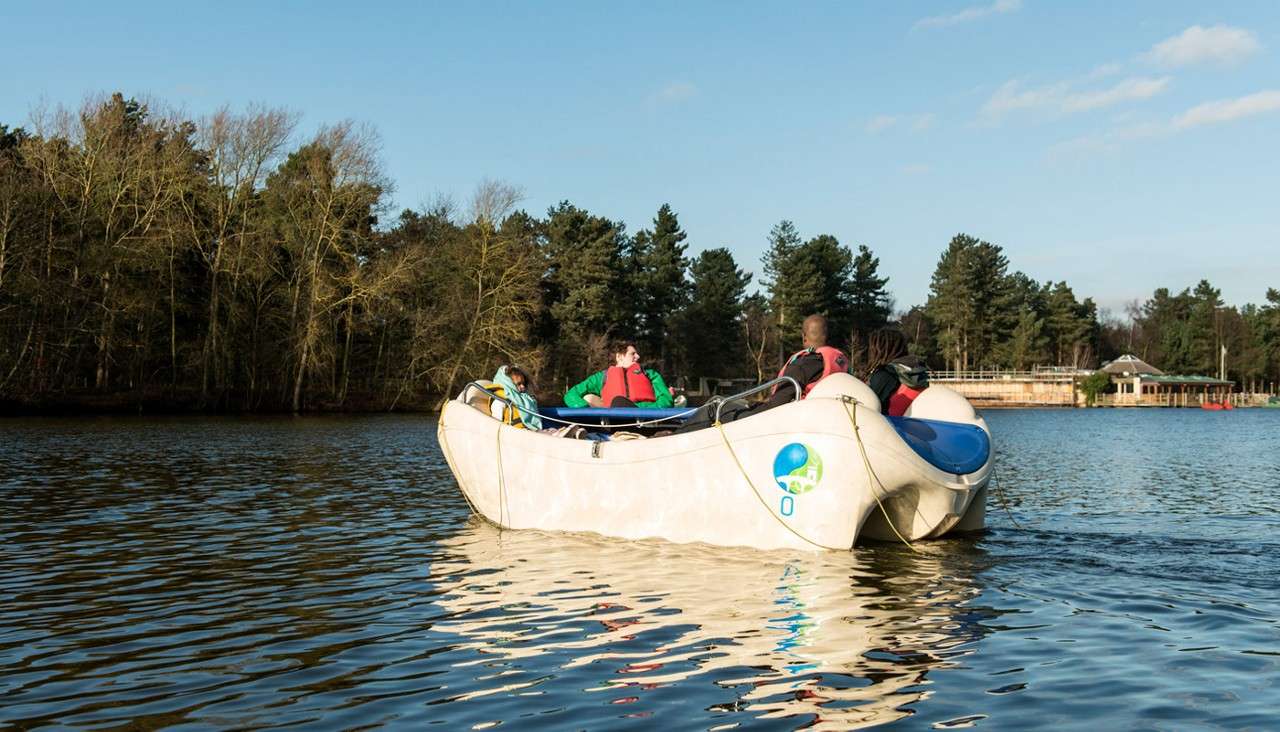 Boat carries several life-jacketed passengers cruising slowly; text on hull: “0”. Context: calm lake with rippling water, forested shoreline, and small lakeside buildings under a clear sky.