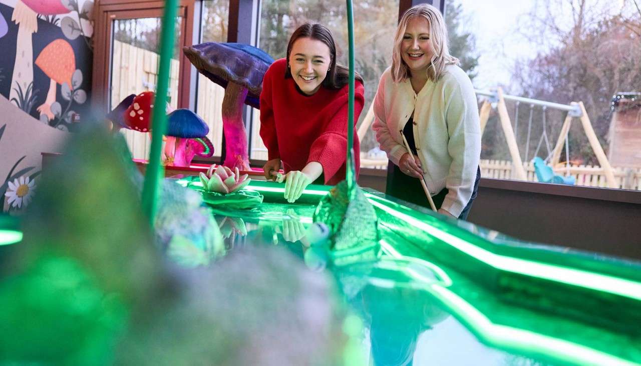 Two women play mini-golf, leaning to aim and smile while guiding the ball. They stand at a neon-green, fantasy-themed course with oversized mushrooms, by windows overlooking a playground and trees.