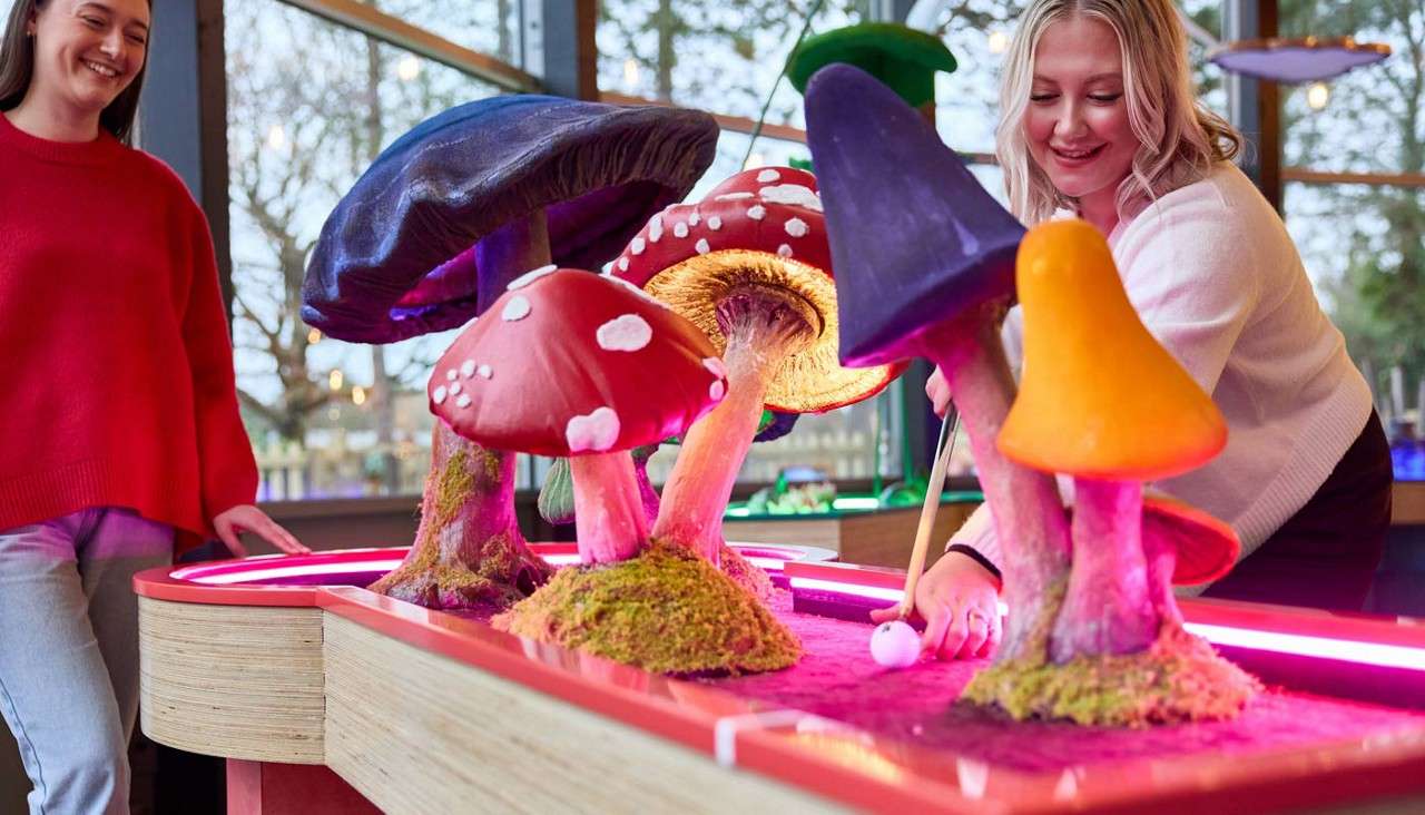 Mini-golf ball rolls as a person putts beneath oversized, colorful mushroom obstacles; another person watches and smiles. Context: indoor neon-lit course beside large windows showing trees outside.