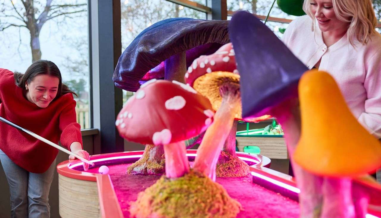Two people putt a white ball, navigating a neon-lit mini-golf hole with oversized colorful mushroom obstacles, inside a windowed venue with trees visible outside.