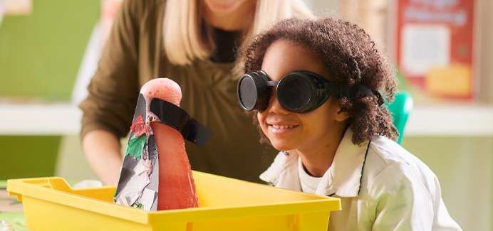 Child wearing safety goggles watches a small homemade volcano bubble with pink foam in a yellow bin, while an adult supervises in a classroom with green walls and posters.