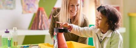 Child touches a homemade volcano model as an adult watches. In a classroom, art projects and supplies surround them; the child wears goggles on their head beside a yellow bin.