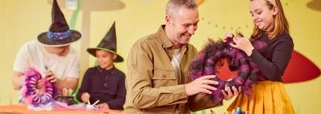 Adults and children craft Halloween decorations, assembling a purple furry spider wreath and gluing materials, while wearing black witch hats in a bright room with tables, markers, scissors, and colorful wall shapes.