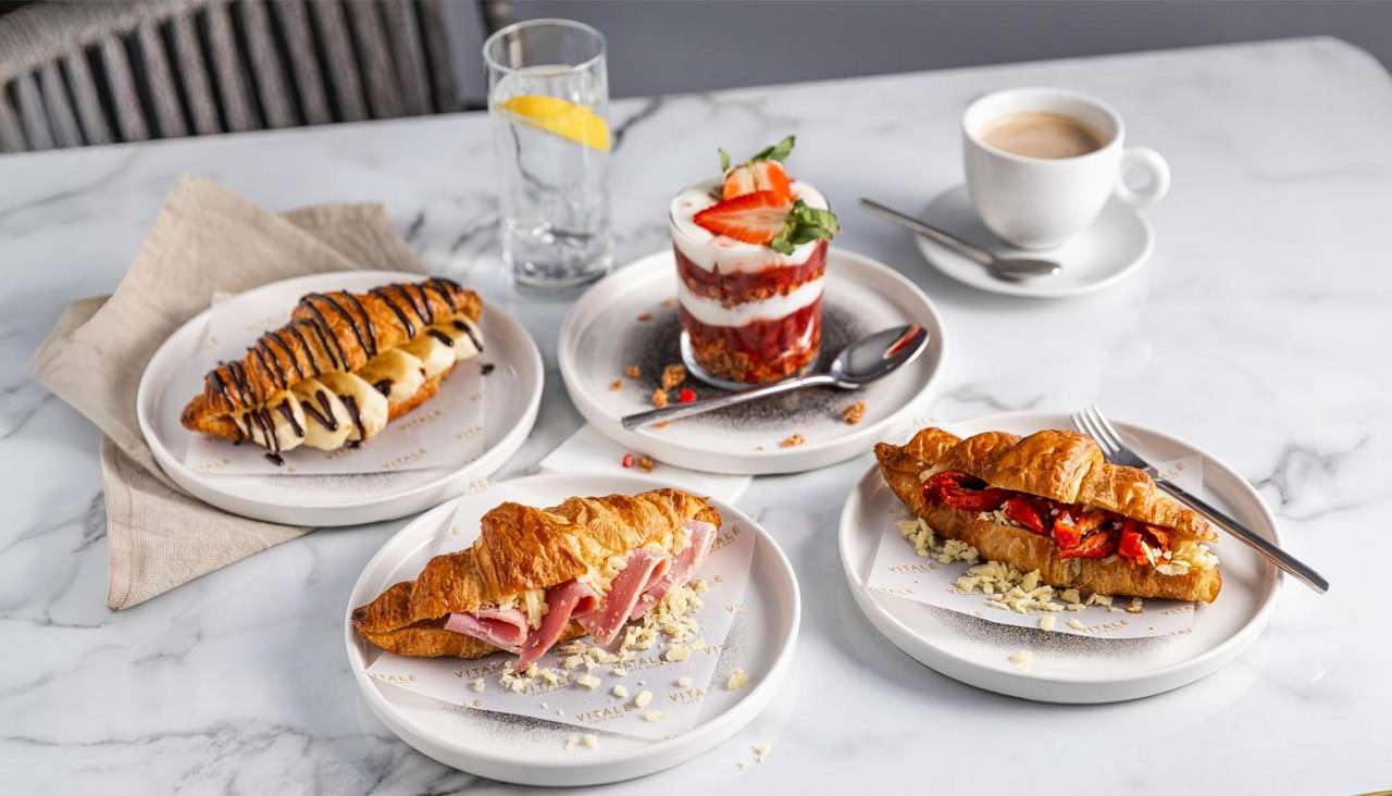 Croissant sandwiches and a chocolate-drizzled pastry sit on plates; a yogurt parfait stands nearby. Context: marble café table with cappuccino, lemon water, cutlery, napkins. Text: VITALE repeated on plate liners.