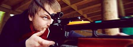 Person aims a scoped rifle, eye to the sight and finger near the trigger; context: wooden-beamed shelter or range with blurred background, suggesting a focused, steady shooting position.