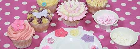 Decorated cupcakes sit on a pink polka-dot tablecloth, surrounded by small bowls and a plate holding pastel fondant flowers and hearts. Frosted cupcakes display floral designs and butterfly toppers.