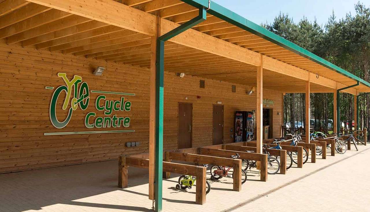 Bicycles line parked bays under a wooden canopy, outside a wood-clad building. Text: Cycle Centre. Vending machines and doors sit by the entrance; trees and parked cars appear in the background.