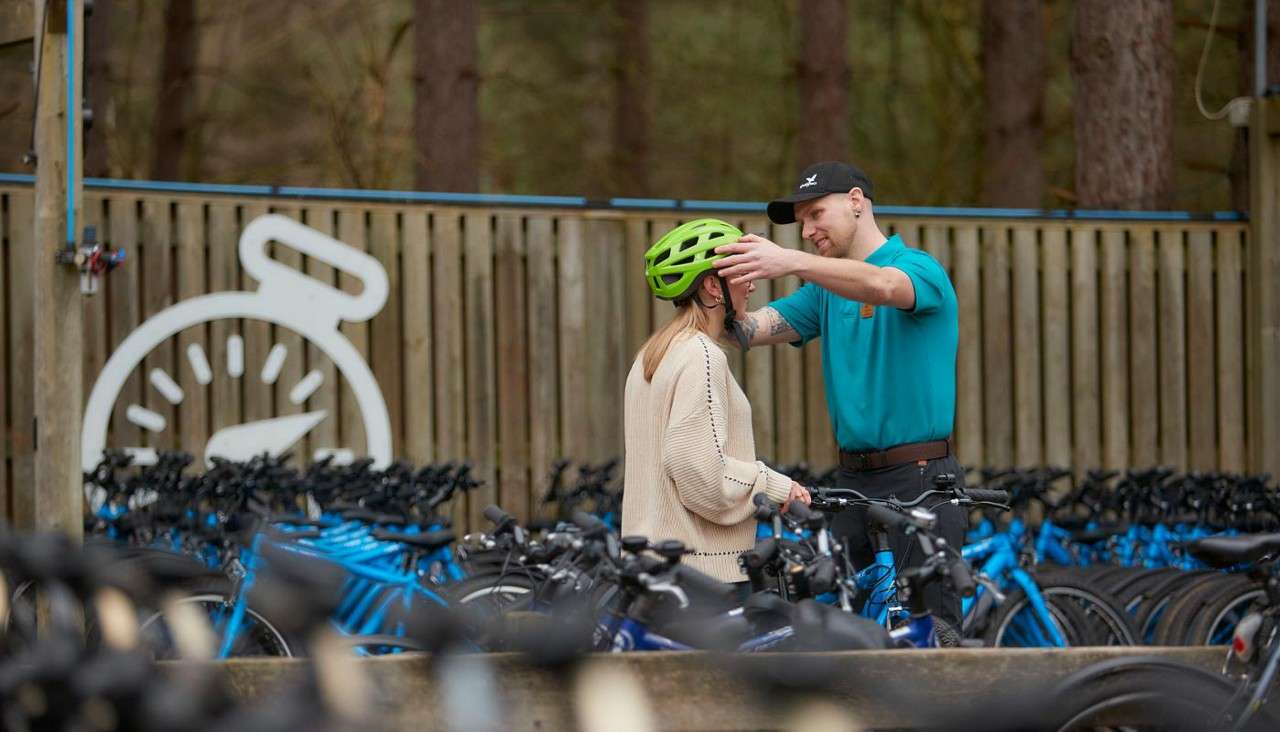 Bike-shop attendant adjusts a woman’s bright green helmet, fitting it securely while she holds a bicycle, amid rows of blue rental bikes near a wooden fence and forest.